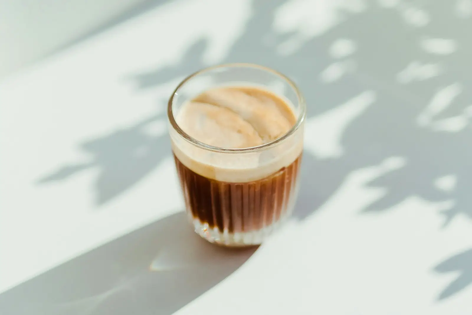 A small glass of cortado on a pale table with leaf shadows
